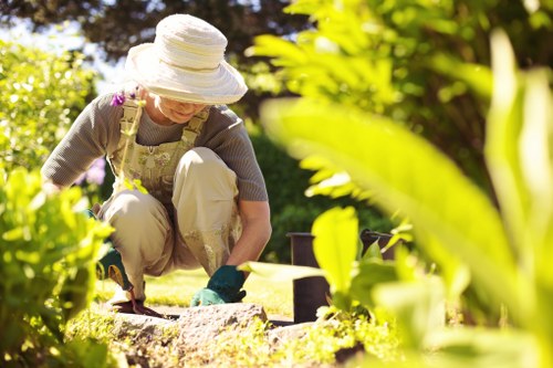 A gardener wearing a wide-brimmed white hat, beige trousers, and a floral apron is crouched down in a lush front garden, engaging in planting or weeding amid well-maintained flower beds and green shrubbery. The garden features a neatly trimmed lawn with dense, vibrant grass, bordered by a variety of leafy plants and flowering shrubs. In the background, there are small garden ornaments or containers, and tall trees provide dappled sunlight reflecting across the scene. The environment appears clean, organized, and reflects regular outdoor maintenance typical of a residential garden in the Elephant and Castle area, with natural tones predominating. The scene is set on a bright day with clear weather, emphasizing the natural beauty and engagement with gardening as part of quality outdoor space management by Landscapers Elephant and Castle, highlighting their expertise in lawn and garden care services.