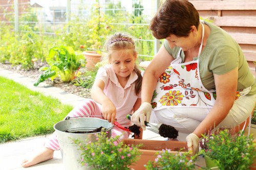 In a well-maintained backyard garden, a woman and a young girl are engaged in planting activity near a flower bed. The garden features a neatly edged lawn with lush, dense grass in the foreground, bordered by a variety of flowering plants and shrubs. Behind them, a fencing area contains green foliage and small trees, indicating a landscaped outdoor space that may be part of a residential property within the Elephant and Castle postcode area. The woman is wearing a floral apron and gardening gloves, gently handling soil and plants in a rectangular planting box, while the girl, dressed in a light pink top and striped trousers, watches attentively. To the side, a metal watering can and additional flower pots are visible, suggesting ongoing garden care. The scene is set under natural daylight, possibly on a mild, overcast day, emphasizing a calm, outdoor gardening environment aligned with professional gardening services offered by Landscapers Elephant and Castle, focused on lawn maintenance, planting, and flower bed improvements in London’s residential gardens. The overall environment appears tidy, with healthy plants ready for cultivation or display, exemplifying an inviting garden scene suitable for supporting localized gardening and landscaping themes.