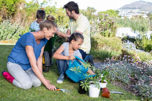 A family engaged in gardening activities in a well-maintained backyard, with a lush green lawn in the foreground and a garden bed filled with flowering plants and shrubs to the right. The mother is kneeling on the grass, planting or tending to a small plant with a gardening fork, while a young girl in a denim jacket is sitting nearby, watering a potted plant with a blue watering can. Behind them, the father and another child are standing and observing, with the father wearing a light-colored shirt and the child wearing a striped top. The garden area includes various gardening tools and pots, with a backdrop of trees and bushes, suggesting a wide, private outdoor space suitable for gardening and lawn care services. Natural daylight illuminates the scene, indicating clear weather conditions, and the overall environment reflects a tidy and vibrant outdoor garden area within a suburban or residential setting in the UK.
