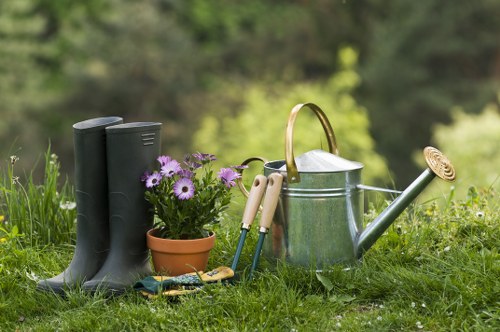 A collection of gardening tools and equipment arranged on a grassy patch in a backyard garden. Visible elements include a pair of black rubber boots, a small terracotta flower pot containing purple and white flowers, a metal watering can with a curved handle and a spout, and a set of small hand tools with wooden handles such as pruning shears and a trowel. The background features blurred green foliage and trees, indicating an outdoor environment with natural light and a warm, possibly sunny day. The scene suggests preparation for planting or garden maintenance, aligning with gardening services offered by Landscapers Elephant and Castle in the local area. This outdoor space has a natural, cared-for appearance with lush grass and a calm, verdant setting typical of a well-maintained front or back garden in the vicinity of London, possibly near Elephant and Castle postcode area.