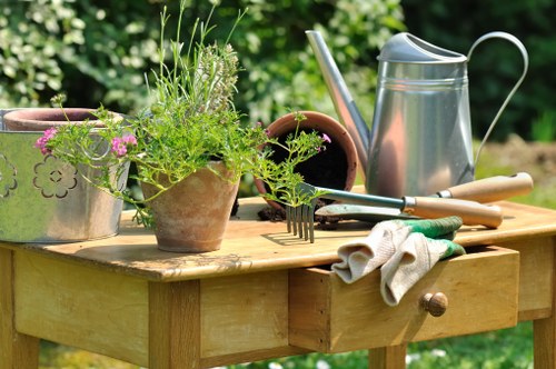 A wooden garden table situated outdoors in a lush green environment, featuring various gardening tools and materials. On the table, there is a terracotta flower pot with a mix of green foliage and flowering plants, alongside several empty terracotta pots. A metal watering can with a long spout sits among the pots, and a small hand rake and garden trowel are resting on the table, with some gloves placed nearby. The background showcases a well-maintained lawn with rich grass and a dense border of trees and shrubs, indicating a landscaped backyard or front garden in the Elephant and Castle area. The scene appears to be set on a sunny day with natural light illuminating the tools and plant containers, supporting professional gardening and landscaping services offered by Landscapers Elephant and Castle.