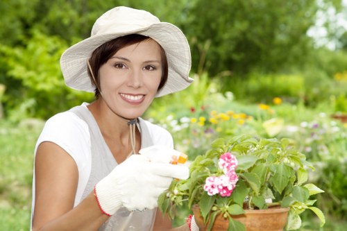 A woman wearing a white wide-brimmed hat, gardening gloves, and a light-colored shirt is tending to a flower bed in a garden with lush green foliage and blooming pink flowers. She is smiling while carefully handling the plants, which are arranged in a container with soil visible around the base. The background shows a well-maintained outdoor space featuring a grassy lawn, a variety of shrubs, and trees, suggesting a landscaped garden area typical of outdoor residential or community spaces in Elephant and Castle. The overcast weather provides soft, even lighting that highlights the natural tones of the plants and the garden environment, supporting gardening and landscaping services by showcasing healthy, vibrant garden features appropriate for both casual and professional outdoor maintenance in London. This scene exemplifies ongoing horticultural care that Landscapers Elephant and Castle provides, emphasizing flower bed creation and garden enhancement in urban outdoor spaces.
