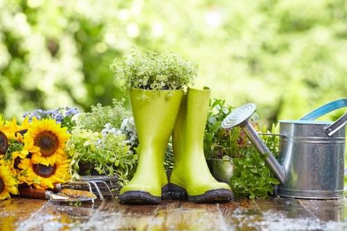 The image depicts a garden scene with a weathered wooden table holding various gardening tools and decorative elements. Prominently, a pair of bright green rubber gardening boots filled with small white flowering plants are placed centrally on the table, suggesting recent planting or decorative arrangement. To the right of the boots, a silver metal watering can with a blue handle is positioned, alongside a smaller silver watering can with a spout. On the left side of the table, a bunch of vibrant yellow sunflowers with dark centers and some purple flowers are displayed, adding colour contrast to the scene. The background features lush, out-of-focus greenery, indicating a well-maintained outdoor garden space, possibly in Elephant and Castle, London. The natural daylight highlights the textures of the wooden surface, the vibrant plant colours, and the metallic watering cans, making this scene reflective of typical gardening tasks and outdoor preparation in a residential garden setting managed by Landscapers Elephant and Castle.