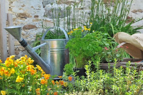 The garden scene features a neatly organized flower bed with a vibrant border of yellow and orange flowering plants at the front, complemented by lush green foliage and small herbaceous plants behind. To the right, a weathered straw hat rests on a garden surface, possibly a stone or paved area. In the center, a metal watering can with a curved handle and long spout is placed among the plants, suggesting ongoing gardening activity. The background includes taller greenery and reeds growing along a stone wall, which provides a rustic backdrop typical of outdoor UK gardens. The lighting appears natural, indicating a bright, possibly sunny day, enhancing the natural tones of the plants, soil, and garden tools. This scene reflects a well-maintained outdoor space, likely part of a residential garden in Elephant and Castle or nearby in central London, designed for gardening and outdoor enjoyment, consistent with the services offered by Landscapers Elephant and Castle. The overall setting emphasizes plant care, garden organisation, and outdoor aesthetic enhancements suitable for local gardening projects including flower bed maintenance and small-scale outdoor improvements.