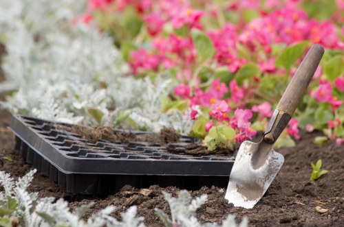A close-up view of a garden bed in a landscaped outdoor space in Elephant and Castle, showing freshly turned soil with a small gardening trowel resting partly in the ground. Surrounding the bed are perennials with white foliage and vibrant pink flowering plants, indicating an early stage of planting or soil preparation for new garden features. In the background, there is a black plastic seed tray filled with soil, suggesting ongoing planting activity. The scene is illuminated by natural daylight under outdoor weather conditions typical of London, with a well-maintained garden area that may be part of a residential or community garden managed by Landscapers Elephant and Castle, highlighting their gardening and landscaping services in the urban outdoor environment.