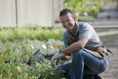 A professional gardener kneeling in a well-maintained outdoor garden area, smiling at the camera while tending to a flower bed with a small gardening fork. The garden features a lush green lawn in the foreground, bordered by a variety of plants, shrubs, and flowering plants, with a wooden fence and trees visible in the background. The soil in the flower bed appears fertile and well-kept, with neat plant arrangements. The scene is lit by natural daylight, suggesting a clear, bright day. The gardener wears a grey shirt, jeans, and a brown apron, indicative of gardening and landscaping services provided by Landscapers Elephant and Castle in the area near South Bank, London, supporting local gardening and outdoor maintenance needs.