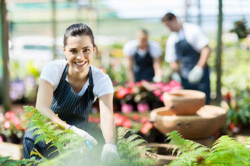 A cheerful woman wearing a striped apron and white gloves is engaged in gardening within a greenhouse or garden centre environment, surrounded by vibrant green ferns and other potted plants. The background features blurred images of other people, possibly staff or visitors, working among flowering plants and garden displays, with sunlight filtering through the glass roof providing bright, natural lighting. The scene showcases a well-maintained outdoor or garden centre setting, with a focus on healthy, lush foliage and a variety of flowering plants arranged on tables and shelves. The woman appears to be tending to the plants, highlighting the cleaning and planting aspects of gardening work. The image captures a professional yet inviting atmosphere, typical of a landscaping or gardening service environment in London or the Elephant and Castle area, emphasizing horticultural care and outdoor maintenance.